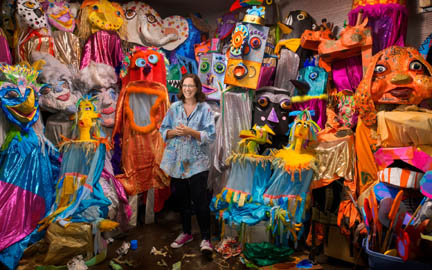 Artist Cheryl Capezzuti, a white woman in a demin shirt with paint splatters on it, stands in her studio surrounded by many colorful, giant puppets.