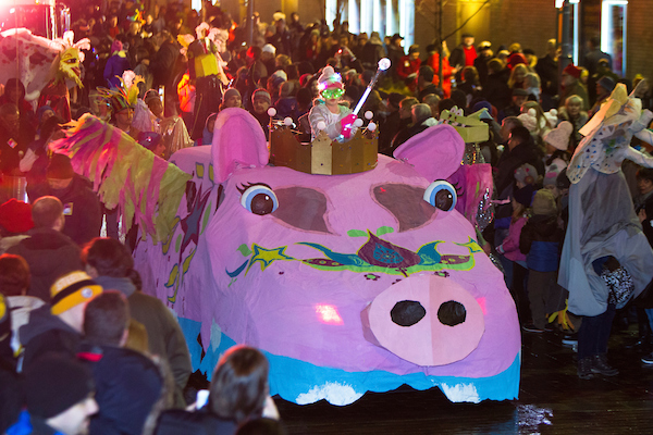 A car that is dressed to look like a giant pig drives through a parade surrounded by a crowd.